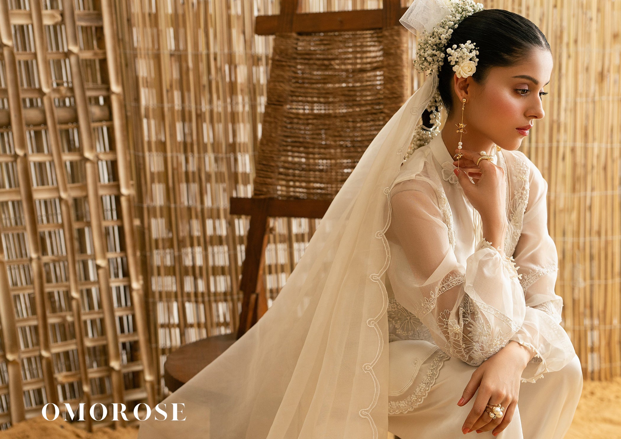 Woman in a white traditional outfit called Aira Mist by Omorose with a veil, sitting against a woven background.