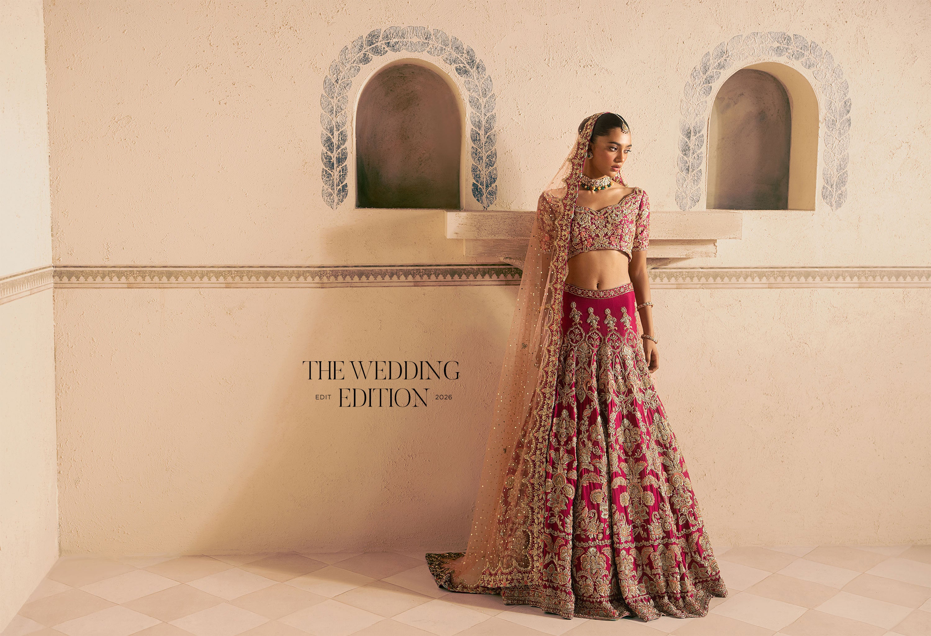 A woman wearing an ornate pink-and-gold hand-embellished Pakistani Indian lehenga choli traditional wedding dress stands in an arched recess. The dress features intricate floral embroidery, a bright pink waistband, and a sheer beige dupatta.