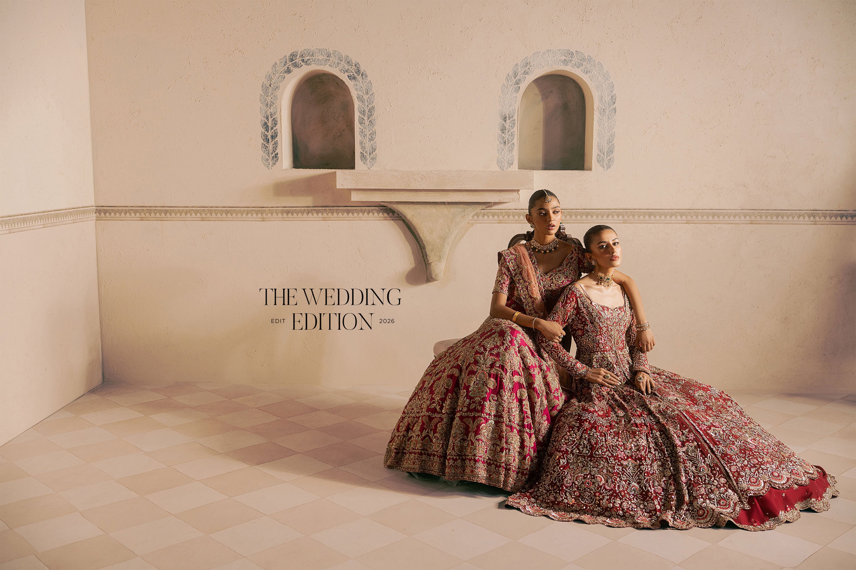 Two women in ornate traditional Indian wedding gowns pose together in a softly lit room. The gowns are heavily hand-embellished in red, pink, and gold tones.